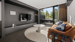 A modern living room with a flat-screen TV, a round coffee table, a leather sofa, and a large sliding glass door leading to an outdoor area. - Stylish Studio w/ Generous Balcony, AC & Parking! (Braddon)