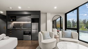 Une chambre d’hôtel moderne dotée d’une petite cuisine, d’un coin salon avec deux canapés, d’une table ronde en marbre et d’une grande fenêtre donnant sur la ville.