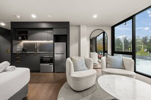 Ein modernes Hotelzimmer mit einer kompakten Küchenzeile, einer Sitzecke mit zwei Sofas, einem runden Marmortisch und einem großen Fenster mit Blick auf die Stadt.