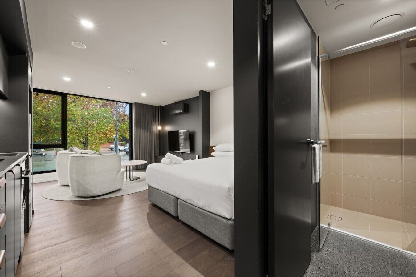 Una moderna camera d'hotel con un letto grande, un'area salotto con una sedia e un tavolo, una televisione e vista sugli alberi.