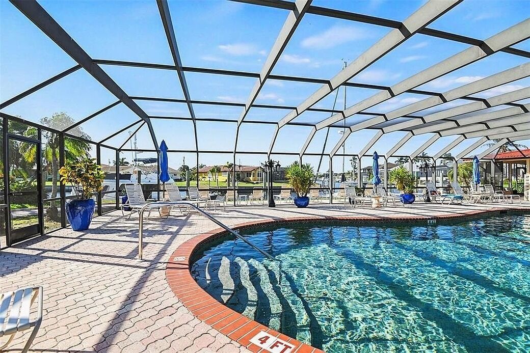 A swimming pool with a glass enclosure, lounge chairs, and potted plants.