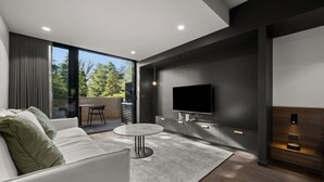 Een moderne woonkamer met een bank, een ronde salontafel, een flatscreen-televisie aan een donkere muur en een balkon met uitzicht op bomen.
