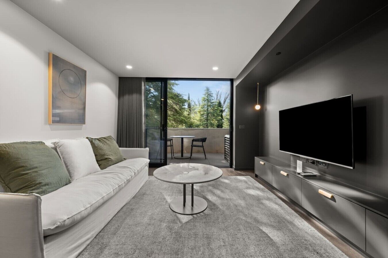 A modern living room with a sofa, a coffee table, a TV, and a balcony with a view of trees.