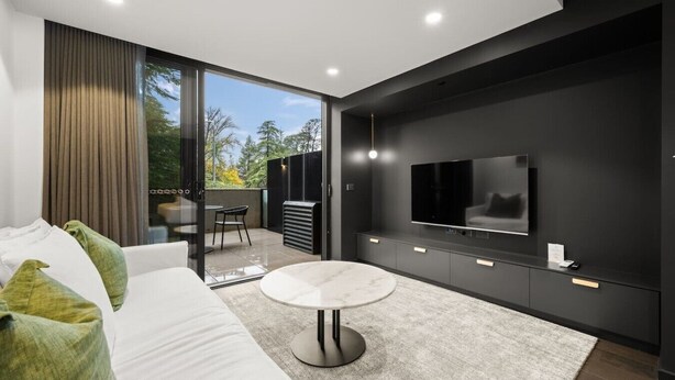 Ein modernes Wohnzimmer mit einer Couch, einem runden Couchtisch, einem an einer dunklen Wand montierten Fernseher und einem Balkon mit Blick auf Bäume.