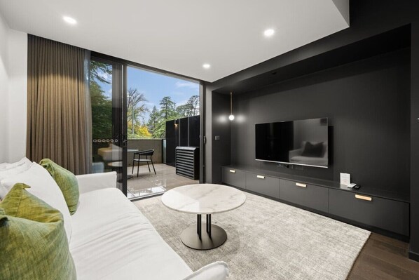 A modern living room with a sofa, a round coffee table, a TV mounted on a dark wall, and a balcony with a view of trees. - Spectacular Studio w/ Haig Park Views & Parking! (Braddon)