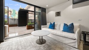 Ein modernes Wohnzimmer mit einem weißen Sofa, blauen Kissen, einem runden Couchtisch und einer Schiebetür zu einem Außenbereich mit Pflanzen.