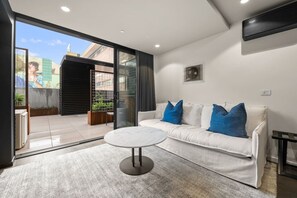 A modern living room with a white sofa, blue cushions, a round coffee table, and a sliding glass door leading to an outdoor area with plants. - Stylish Canberra Studio with BBQ & Terrace (Braddon)