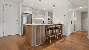 A modern kitchen with a central island, wooden chairs, and a stainless steel refrigerator. - 2BR Haig Park View w/ Balcony & Secure Car Space! (Braddon)
