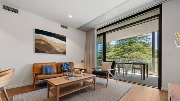 A modern living room with a sofa, coffee table, and chairs, a painting on the wall, and a balcony with outdoor furniture. - 2BR Haig Park View w/ Balcony & Secure Car Space! (Braddon)