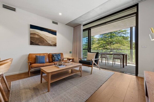 Ein modernes Wohnzimmer mit einer Couch, einem Couchtisch und Sesseln, einem Gemälde an der Wand und einem Balkon mit Gartenmöbeln.