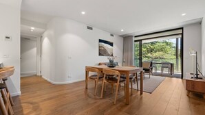 A spacious room with a wooden dining table, chairs, and a large window offering a view of greenery. - 2BR Haig Park View w/ Balcony & Secure Car Space! (Braddon)
