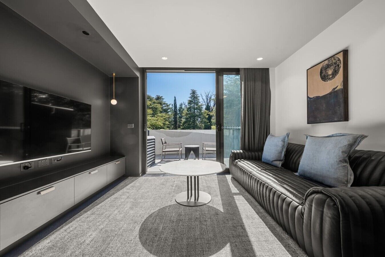 A modern living room with a flat-screen TV, a leather sofa, a round coffee table, and a balcony with outdoor furniture.