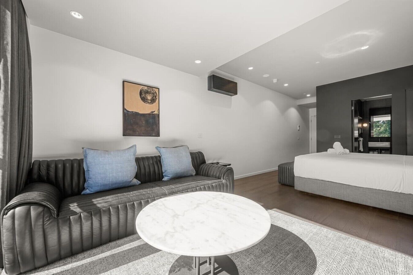 Una habitación de hotel moderna con un sofá gris, una mesa redonda de mármol, una cama y un cuadro en la pared.