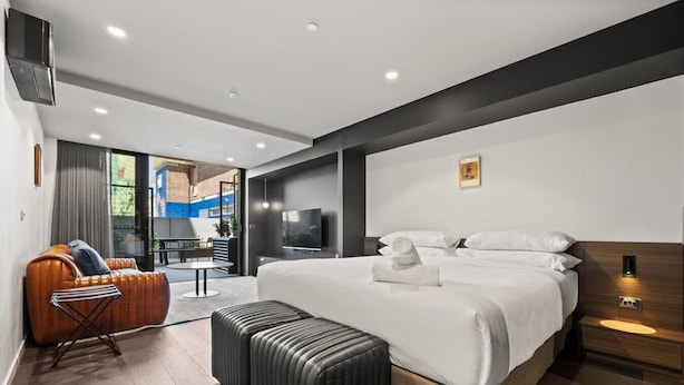 Une chambre d’hôtel moderne dotée d’un grand lit, d’un coin salon avec un canapé et un fauteuil, d’une télévision et offrant une vue sur l’extérieur.
