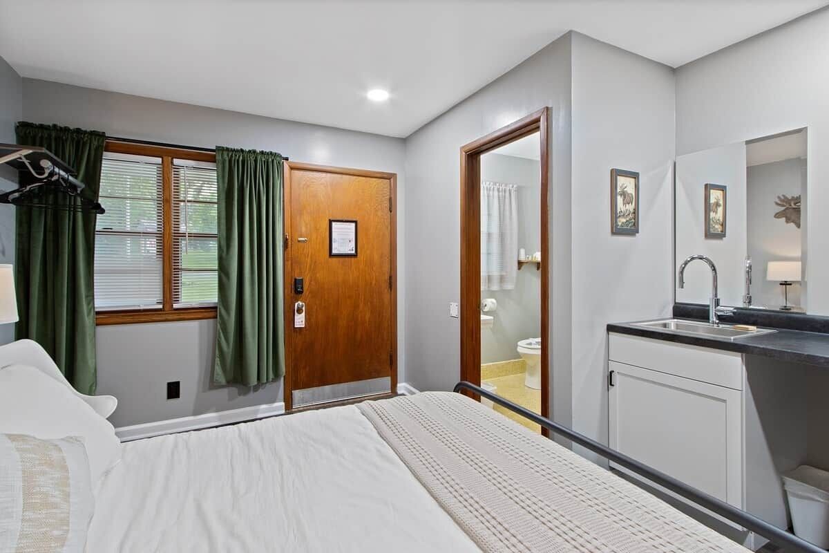 A bedroom with a bed, a wooden door, a window with curtains, a bathroom with a toilet, and a sink.