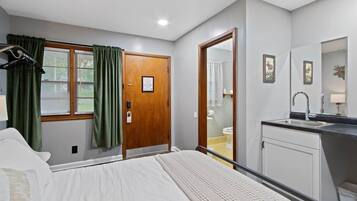 A bedroom with a bed, a wooden door, a window with curtains, a bathroom with a toilet, and a sink.