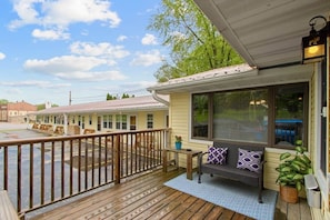 A wooden deck with a bench and potted plants, a covered porch with a window, and a building in the background.