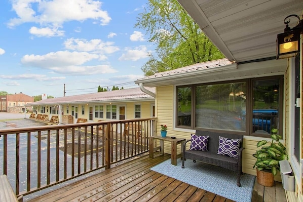 A wooden deck with a bench and potted plants, a covered porch with a window, and a building in the background.