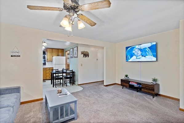 Un salon avec un ventilateur de plafond, un téléviseur fixé au mur, un coin repas avec des chaises et une table, et une cuisine à l’arrière-plan.