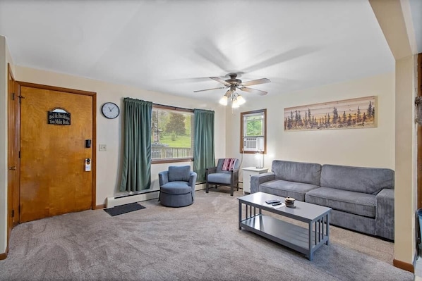 A living room with a ceiling fan, a sofa, two armchairs, a coffee table, a clock, and a door with a sign.