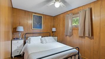 Un dormitorio con una cama, dos mesitas de noche, un ventilador de techo, una ventana con cortinas y un cuadro colgado en la pared.