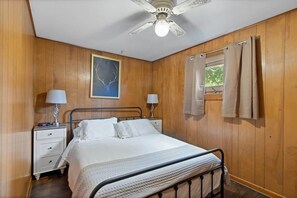 Une chambre à coucher comprenant un lit, deux tables de chevet, un ventilateur de plafond, une fenêtre avec des rideaux et un tableau accroché au mur.