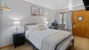 A bedroom with a bed, bedside tables, a desk with a chair, a window with curtains, and two framed pictures on the wall. - Room 5, Kozy Rest Motel (Montello)