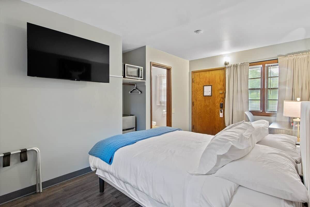 A bedroom with a bed, a TV mounted on the wall, a microwave, and a door leading to another room.