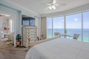 Ein Schlafzimmer mit einem großen Bett, einem Fernseher, einem Schminktisch, einem Sessel, einem kleinen Tisch mit einer Pflanze und einem Balkon mit Meerblick.