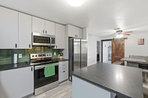 Una cocina moderna con electrodomésticos de acero inoxidable, microondas, horno y refrigerador. Hay un comedor con mesa de madera y bancos.