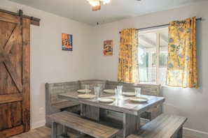 Un comedor con un banco de madera, una mesa con platos y vasos, y un ventanal con cortinas.