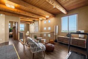 Ein geräumiges Zimmer mit Holzbalkendecke, einer Treppe, einer Sitzecke mit einem gemusterten Sessel, einem runden Hocker und Blick durch ein Fenster.