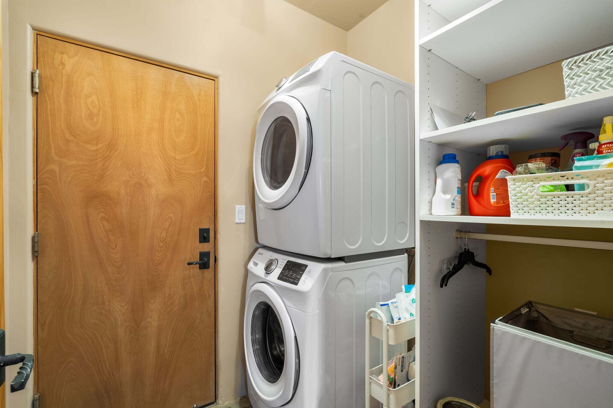 A laundry room with stacked washers and dryers, a wooden door, and shelves with cleaning supplies.