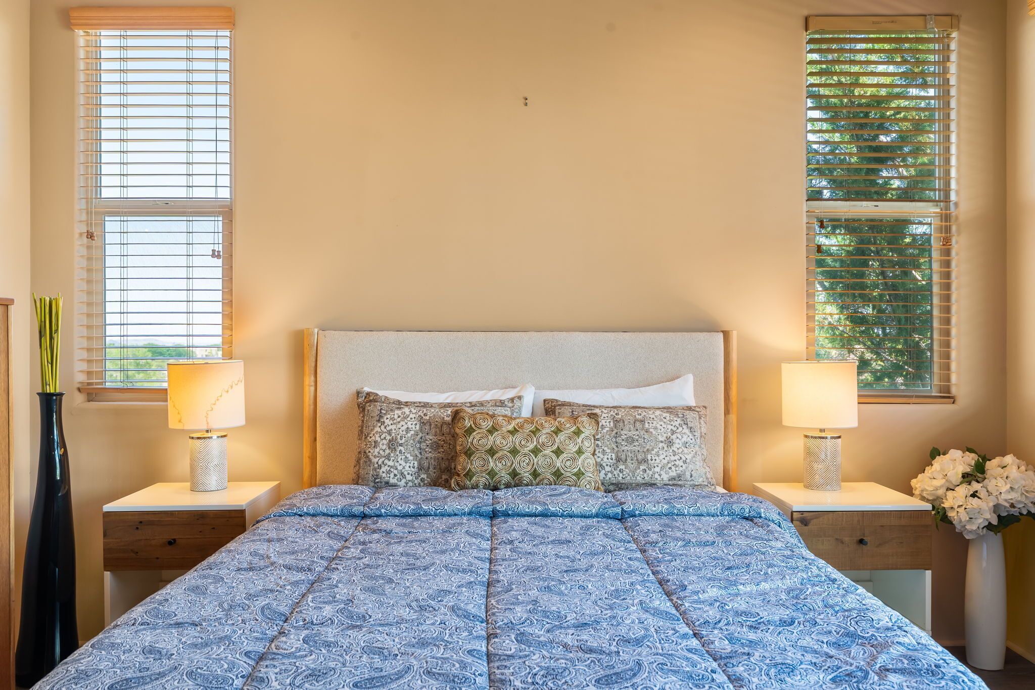 Une chambre à coucher avec un lit, deux lampes de chevet, une table de chevet, un vase avec des fleurs et deux fenêtres équipées de stores.