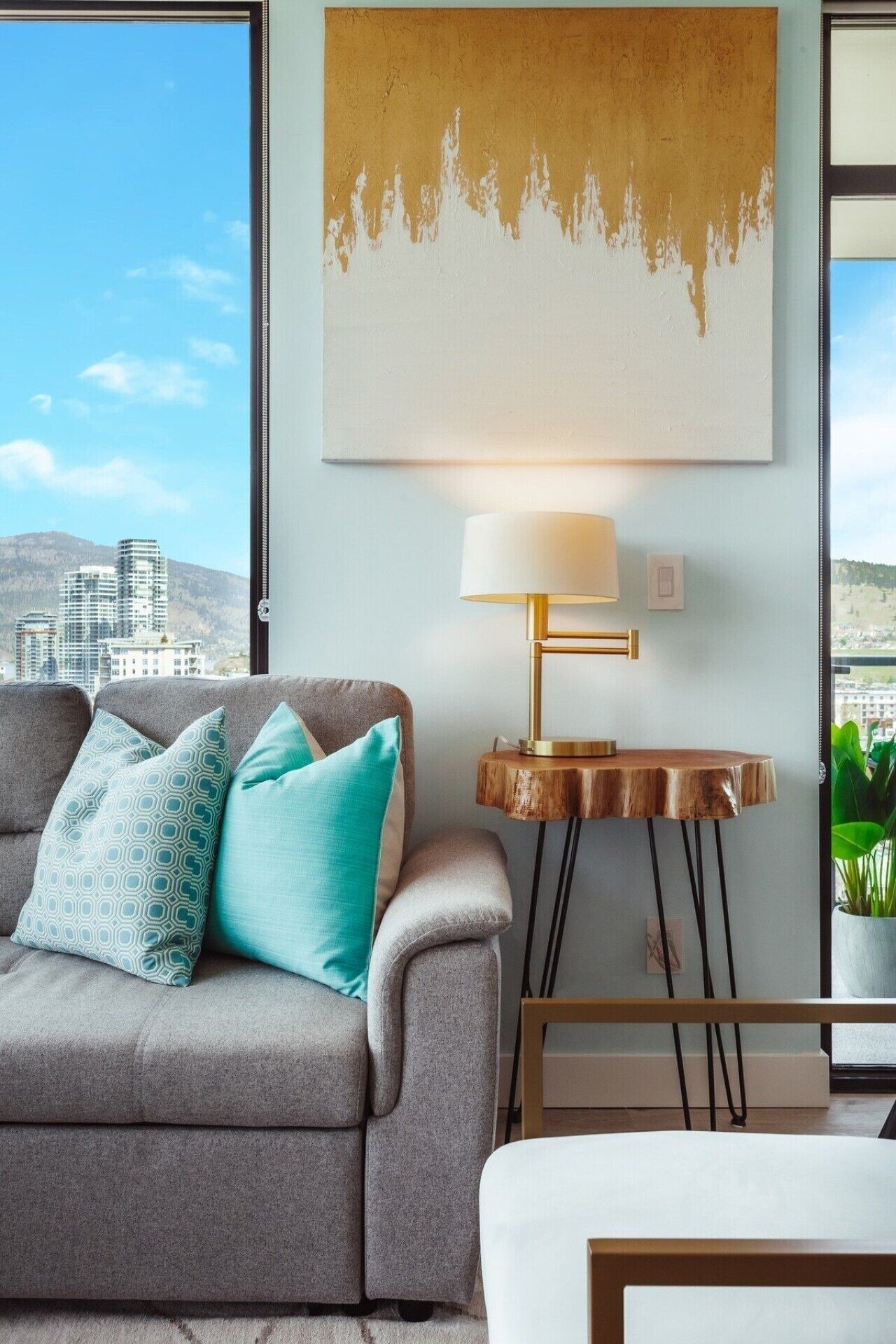 Een moderne woonkamer met een grijze bank, kussens in teal-tint, een houten bijzettafeltje met een lamp, en een stadsgezicht door grote ramen.