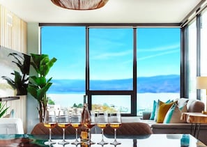 A modern living room with a large window, a dining table with glasses of wine, and a sofa with cushions.
