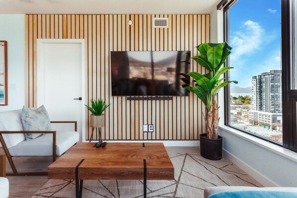 A modern living room with a TV mounted on a wooden panel wall, a sofa, a coffee table, and a large window with a city view.