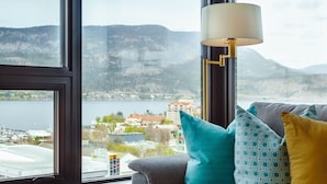 A modern living room with a grey sofa, a blue throw blanket, and a lamp. A large window offers a view of a lake and mountains.