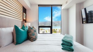 Ett hotellrum med en säng, kuddar, utsikt över en vattenyta och ett stort fönster.