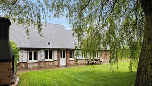 A brick building with white shutters, a picnic table, and a tree in the foreground. - Gîtes de France® - The Little House on the Prairie (ANCRETTEVILLE SUR MER)