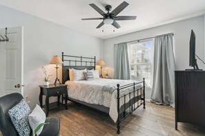 A bedroom with a bed, bedside tables, a chair, a desk, a ceiling fan, and a window with curtains.