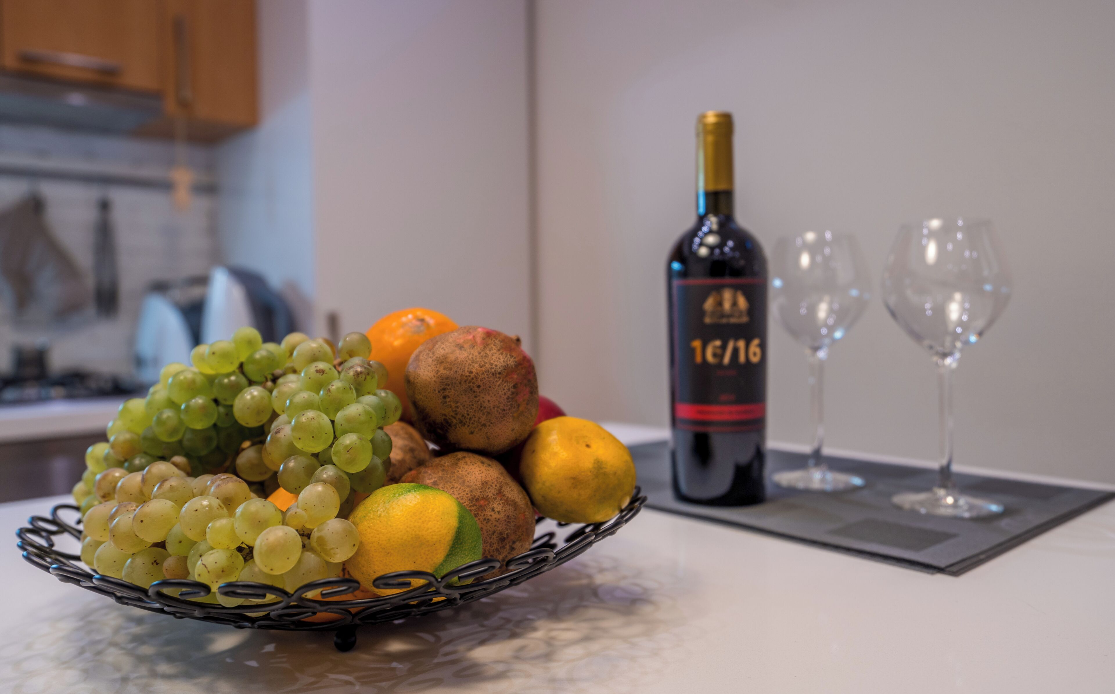 Una ciotola di frutta con uva, un’arancia e un kiwi su un piano di cucina, una bottiglia di vino e due bicchieri da vino.