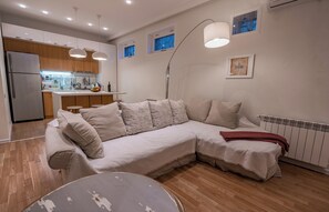 Een moderne woonkamer met een hoekbank, een eethoek met een keuken op de achtergrond, hanglampen en een radiator.
