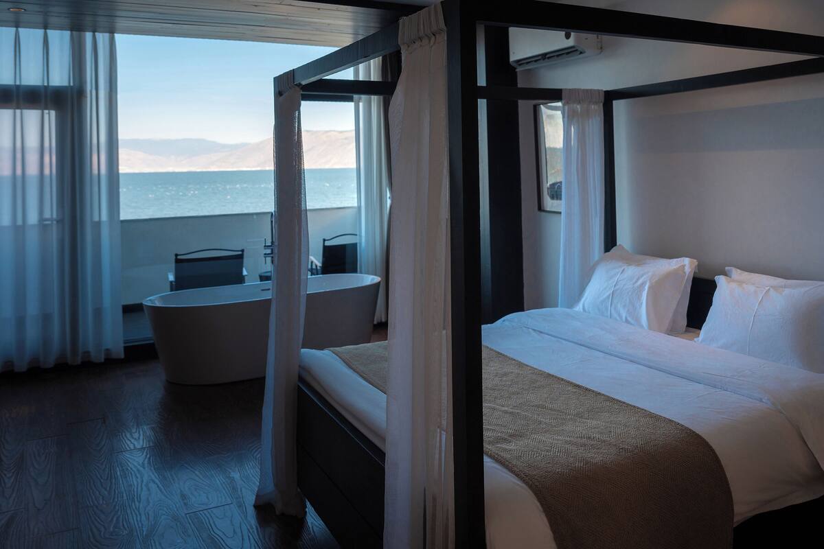 A four-poster bed with white linens, a round table, and a view of the sea.