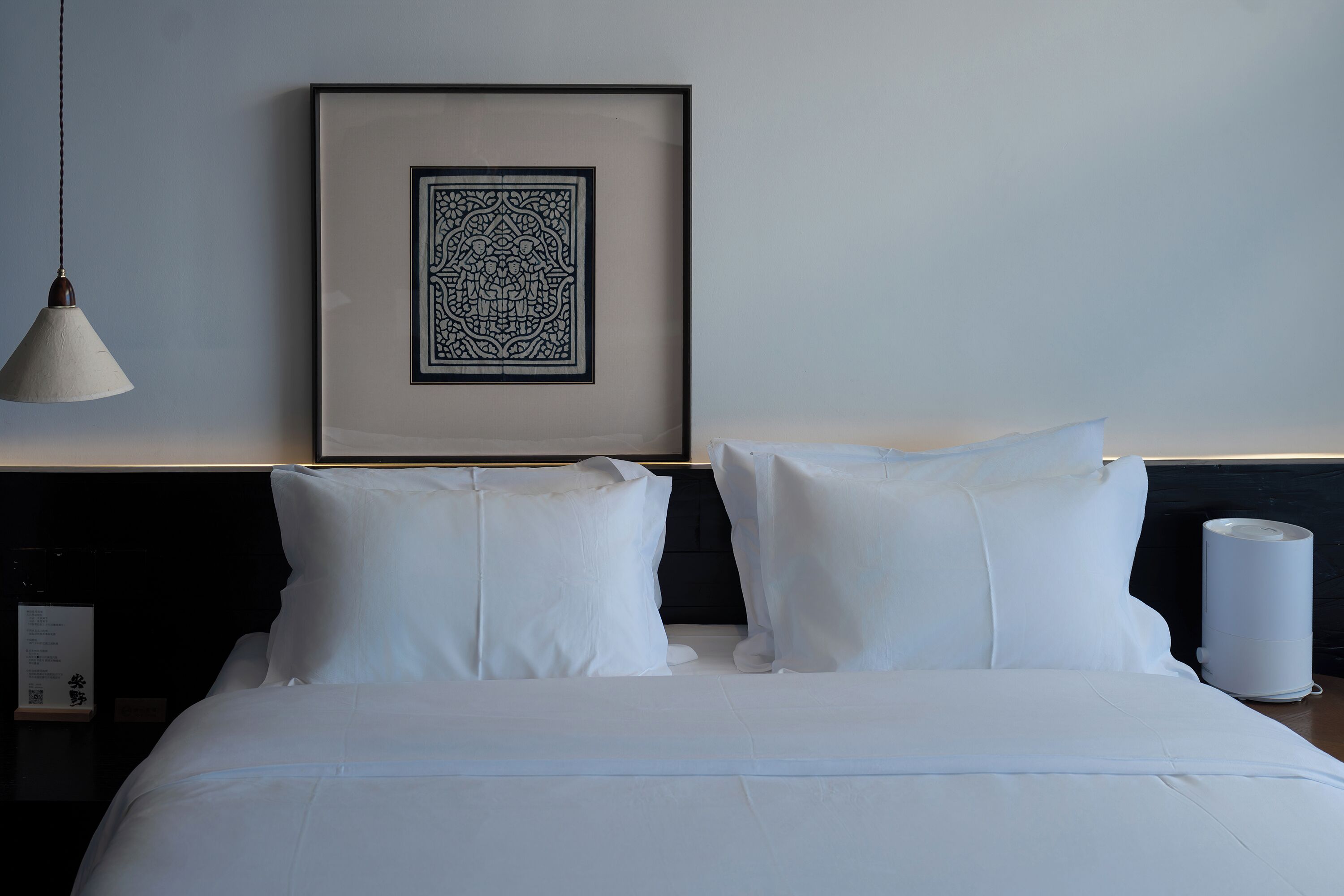 A neatly made bed with white linens, a framed artwork on the wall, and a bedside lamp.