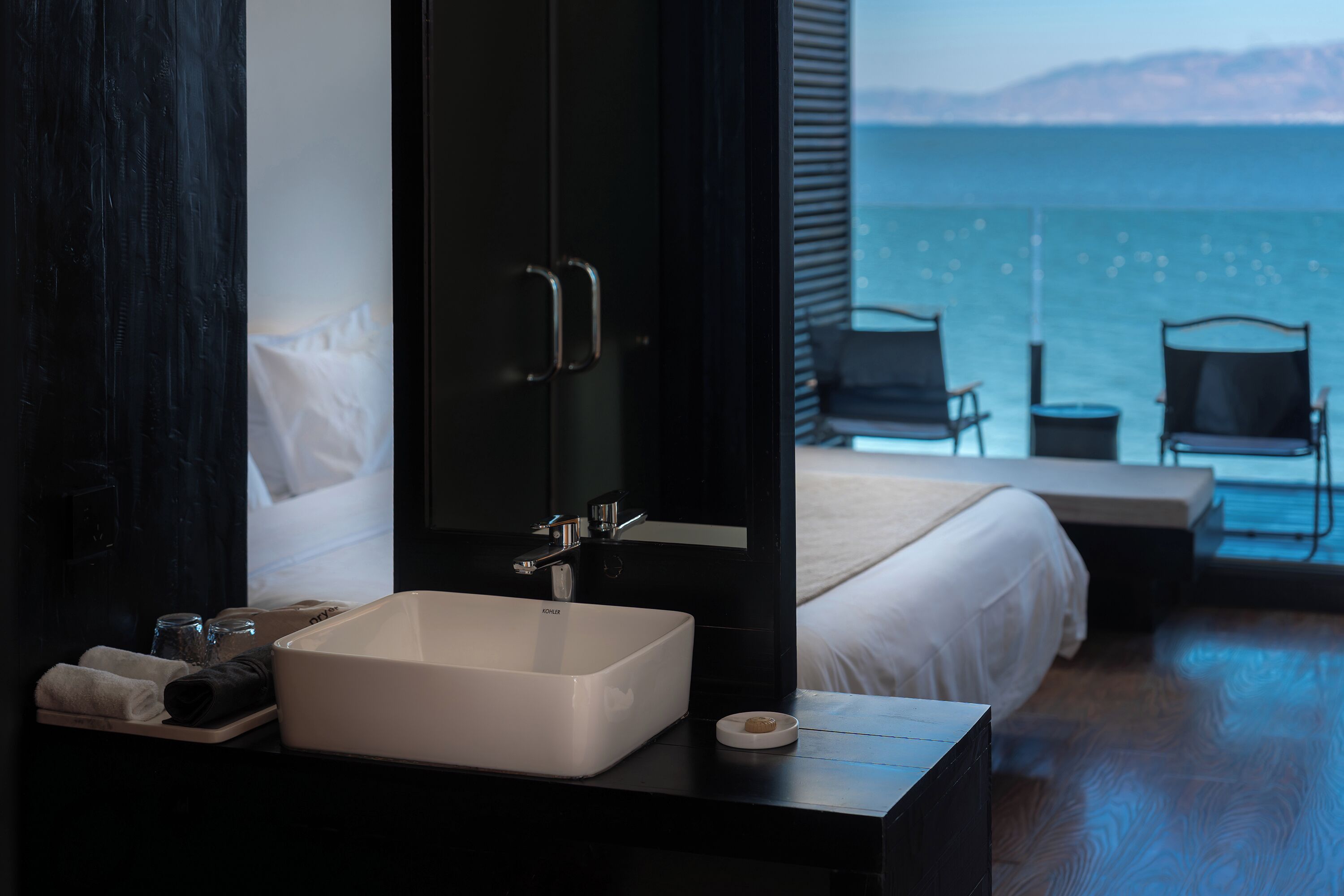 Habitación de hotel con un ventanal grande, una cama, una mesita de noche con lavamanos y vistas al mar.
