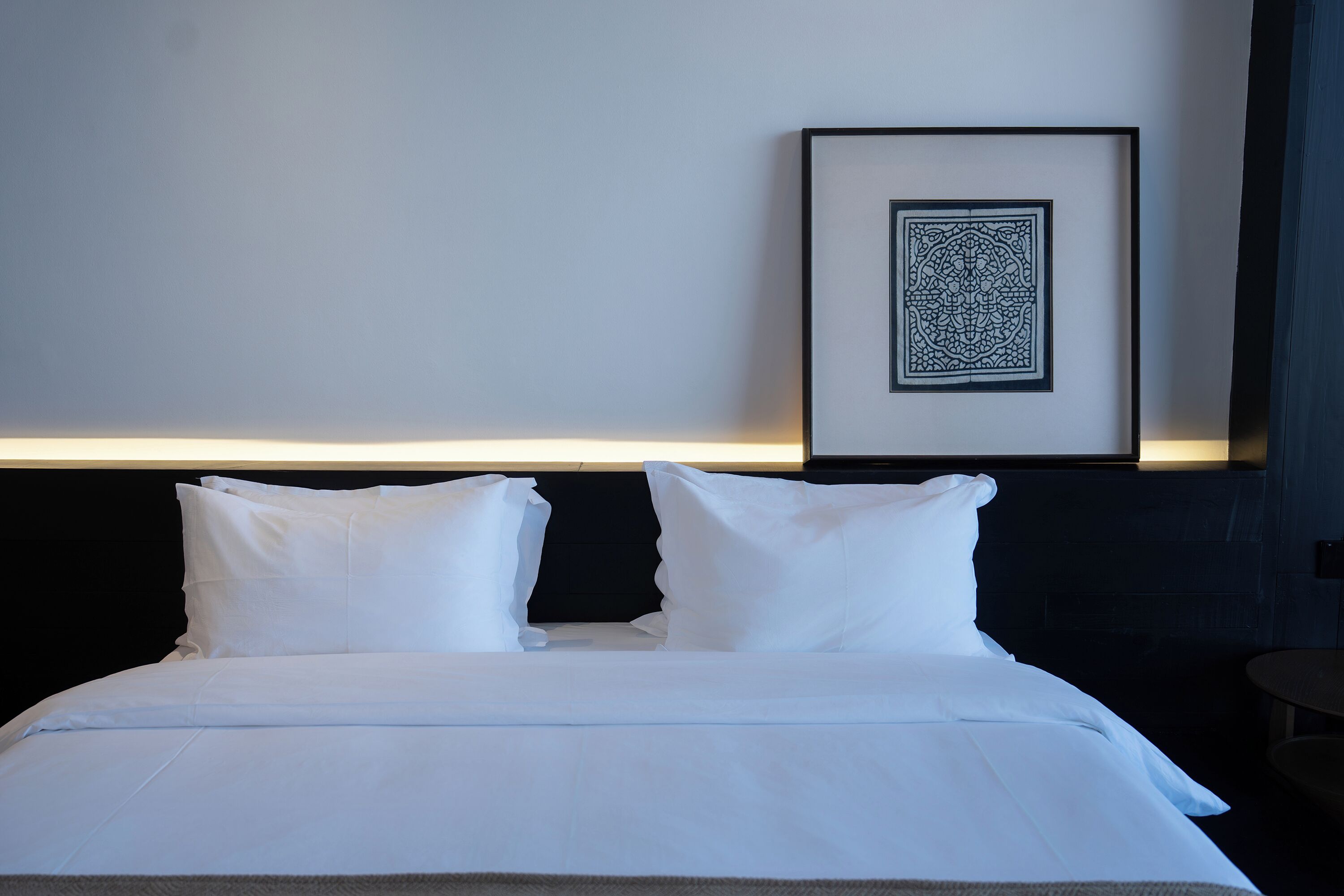 Un lit bien fait, avec du linge de lit blanc, une œuvre d’art encadrée accrochée au mur et une petite table ronde placée à côté du lit.