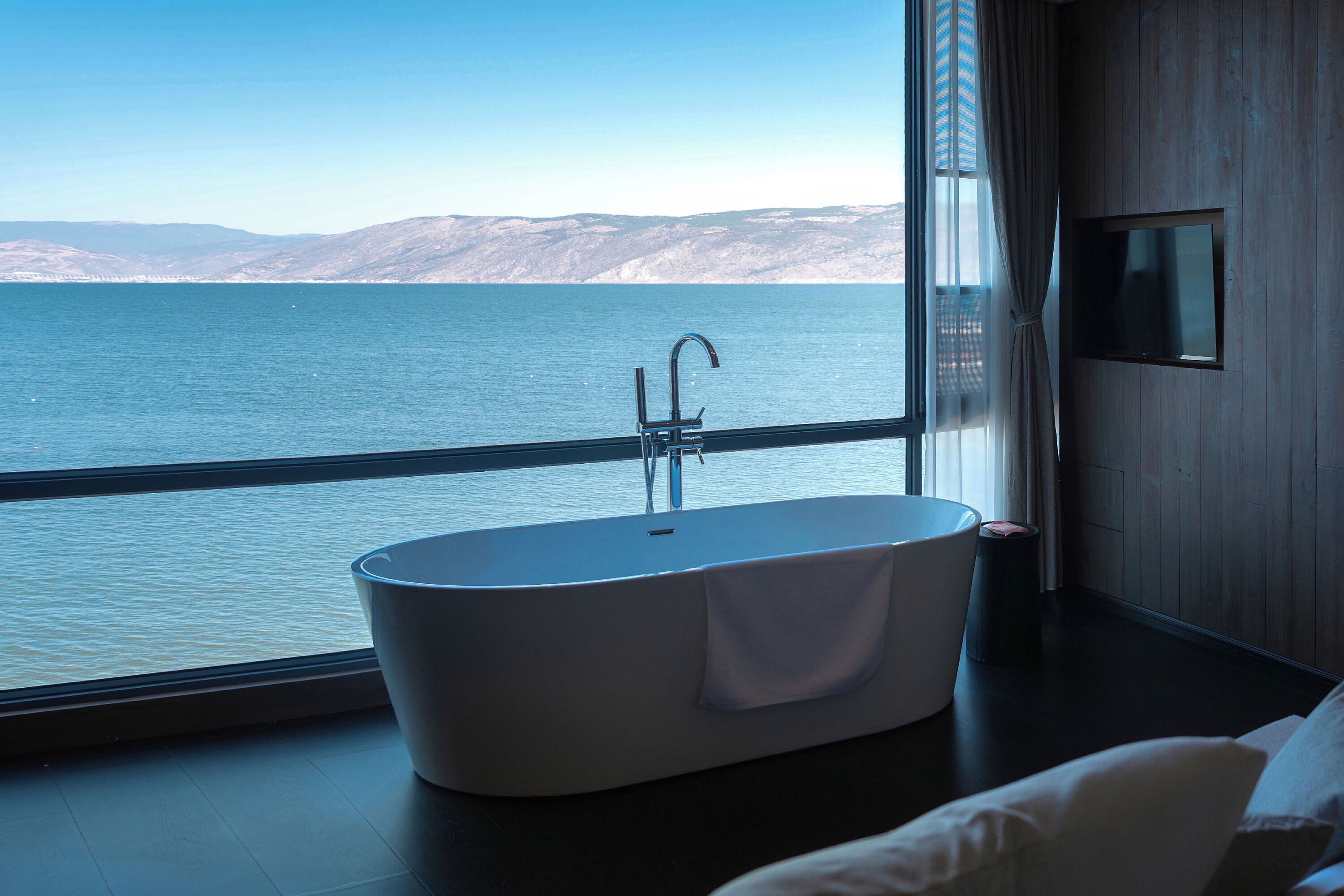 A modern bathroom with a freestanding bathtub, a large window with a view of a body of water and distant mountains, and a flat-screen TV mounted on the wall.