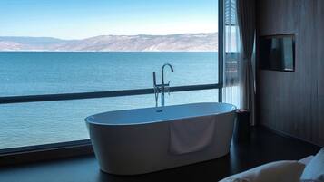 A modern bathroom with a freestanding bathtub, a large window with a view of a body of water and distant mountains, and a flat-screen TV mounted on the wall.