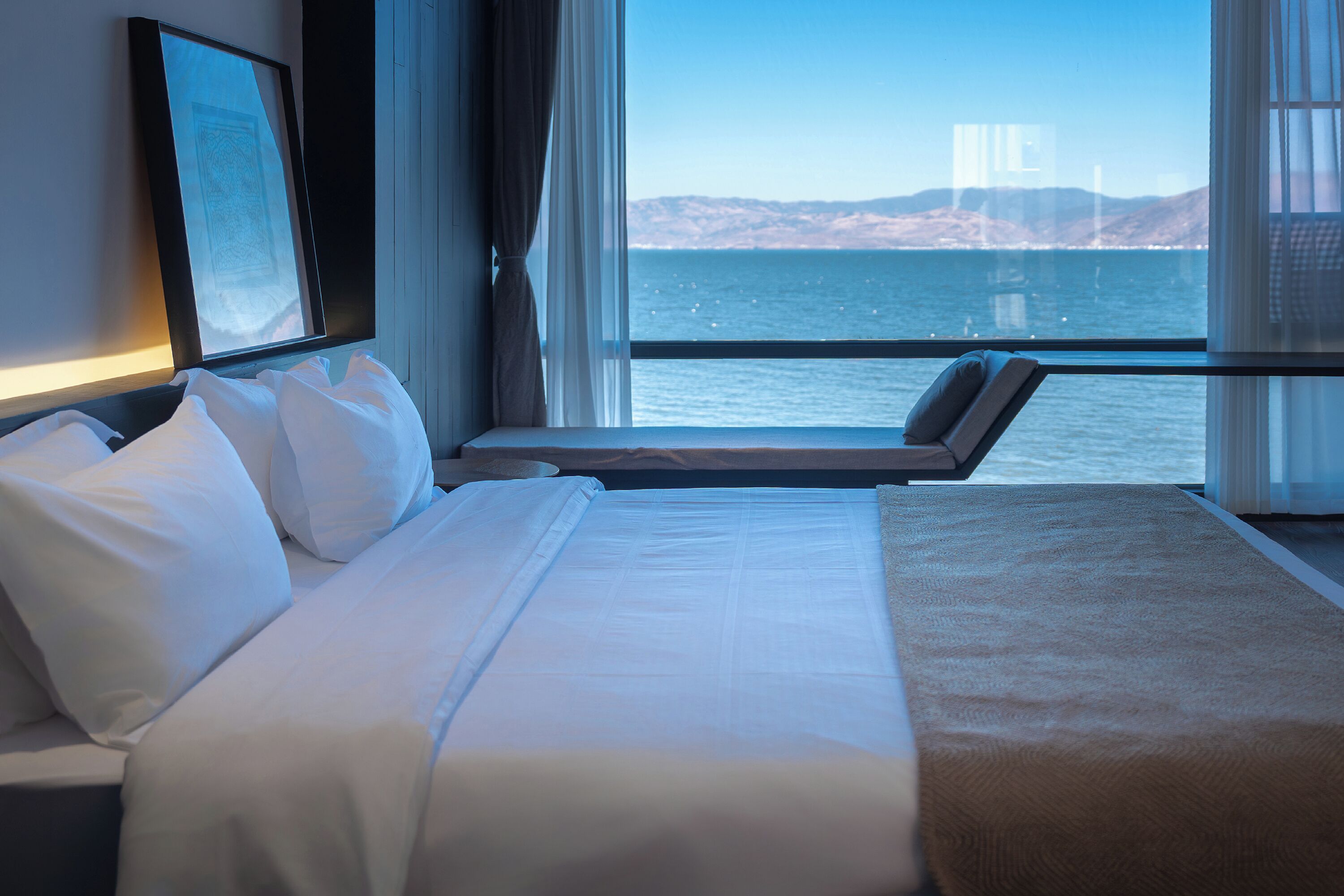 A hotel room with a bed, a window with a view of the sea and a bridge, and a framed picture on the wall.
